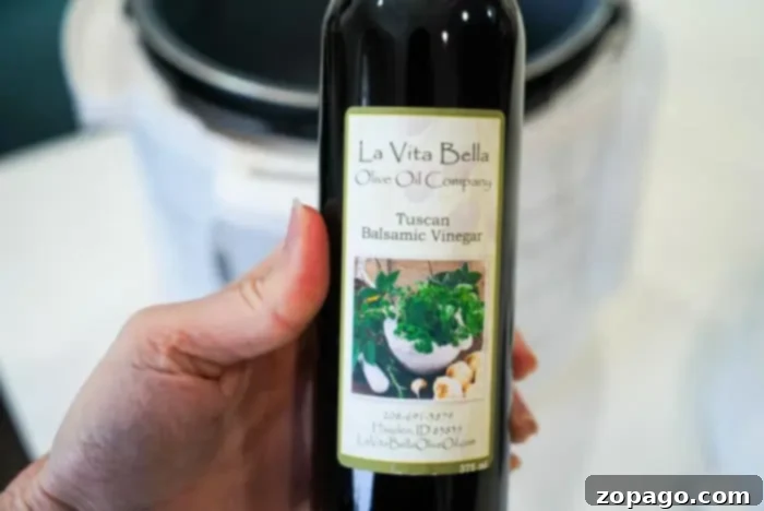 Perfect Beef Arm Roast 6 A hand holds a bottle of La Vita Bella Olive Oil Company's Tuscan Balsamic Vinegar, perfect for enhancing your beef arm roast recipe. The label features images of herbs and garlic, beautifully showcased against a blurred background.