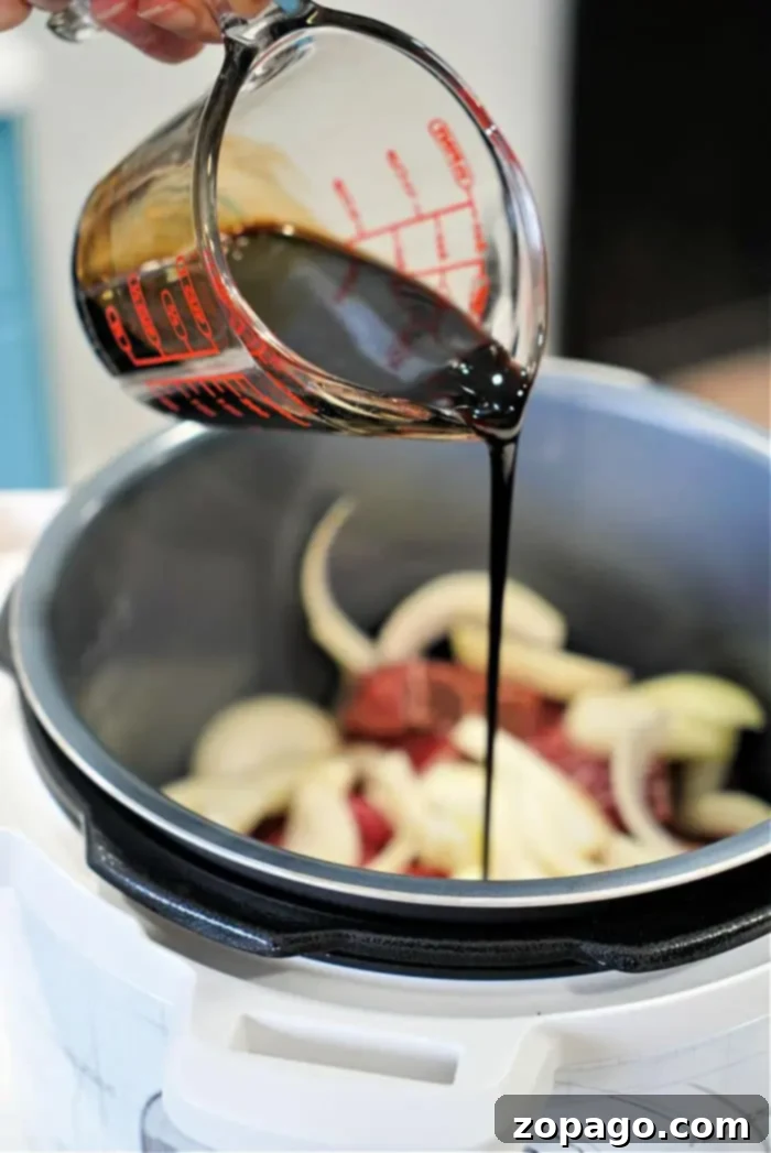 Perfect Beef Arm Roast 7 A hand is pouring a balsamic vinegar from a measuring cup into an electric cooker containing sliced onions and beef arm roast. The cooker's lid is open, spotlighting the ingredients as they meld into a mouthwatering recipe.
