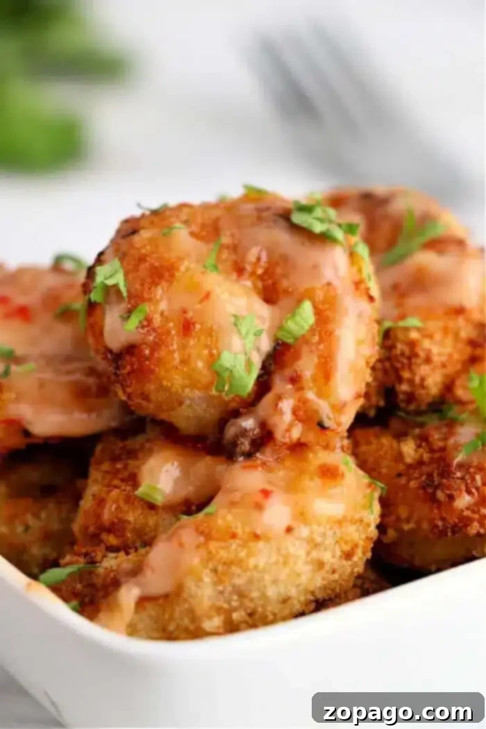 Crispy Air-Fried Bang Bang Shrimp 2 A close-up shot of a white dish filled with air fryer bang bang shrimp, topped with a drizzle of pink sauce and garnished with chopped green herbs. A softly blurred fork and greenery are visible in the background.
