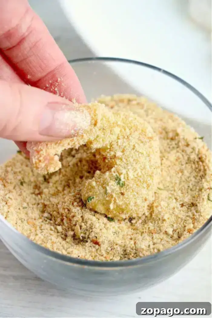 Crispy Air-Fried Bang Bang Shrimp 8 A hand is shown dipping a piece of bang bang shrimp, into a bowl of seasoned bread crumbs. The bread crumbs appear coarse and contain various herbs. The partly coated shrimp will soon be ready for the air fryer, promising a crispy and flavorful delight.