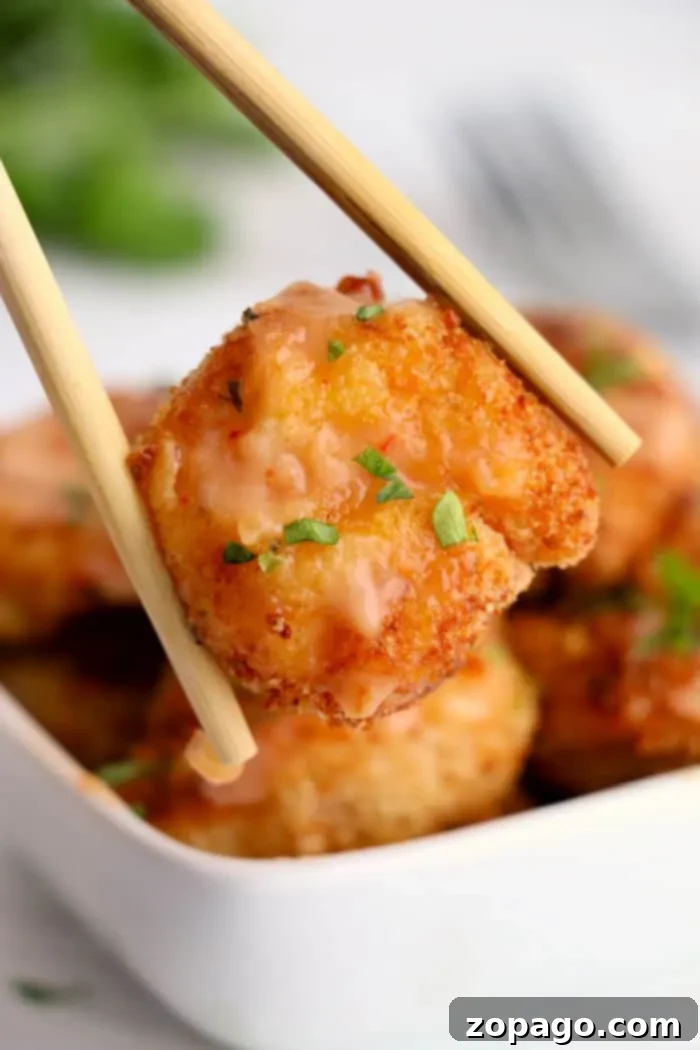 Crispy Air-Fried Bang Bang Shrimp 10 A close-up of chopsticks holding a piece of breaded and fried shrimp, garnished with chopped herbs and a light sauce that gives it an almost bang bang shrimp vibe. In the background, there is a square white bowl filled with more air-fried pieces of the same delicious breaded tofu.