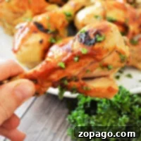 Effortless Instant Pot Frozen Chicken Drumsticks 9 A close-up of a hand holding a grilled chicken drumstick garnished with parsley reveals juicy goodness, reminiscent of flavors unlocked from frozen chicken legs when seamlessly cooked in an instant pot. More drumsticks grace a plate in the background, surrounded by fresh parsley on the wooden surface.