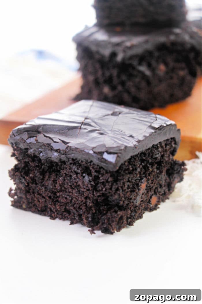 Hidden Veggie Brownies 5 A close-up of a piece of chocolate cake with glossy, dark chocolate frosting, reminiscent of a rich zucchini brownie recipe, is placed on a white surface. Another piece of cake is visible in the blurred background on a wooden board.