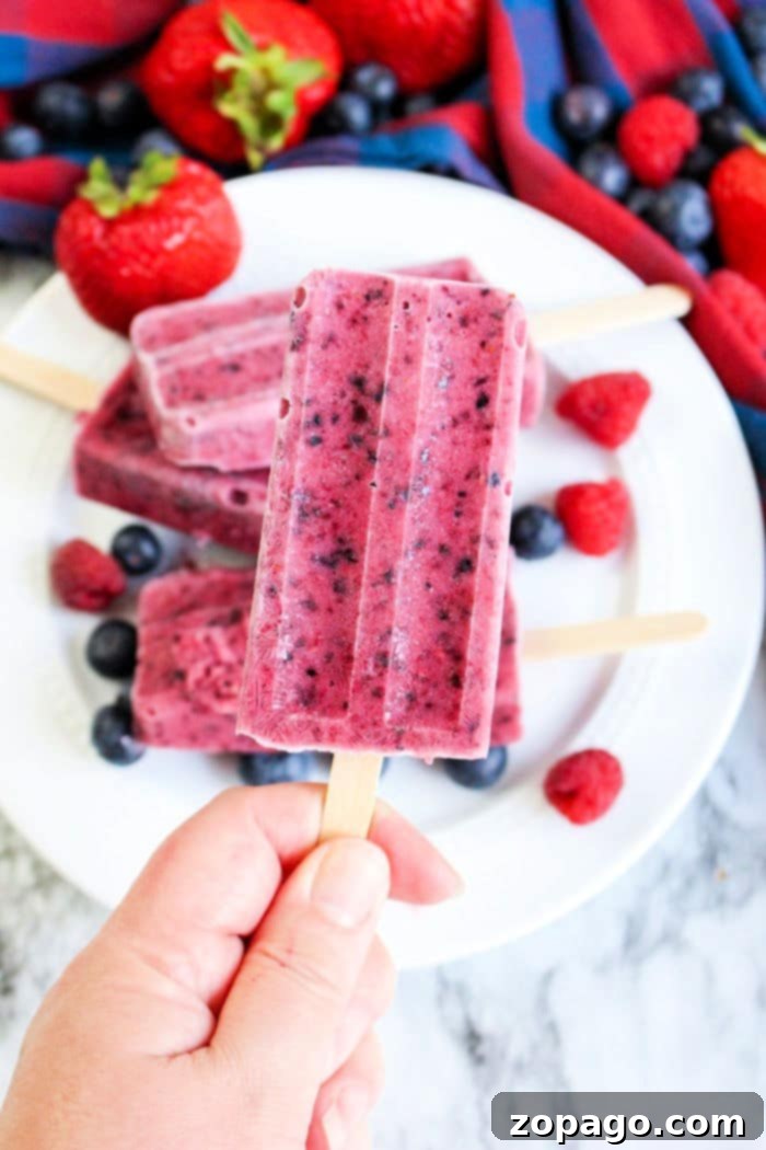 Create Perfect Homemade Popsicles 2 A hand holds a berry popsicle, showcasing a vibrant treat among more popsicles and fresh berries on a plate—an enticing glimpse into the delightful world of how to make homemade popsicles.