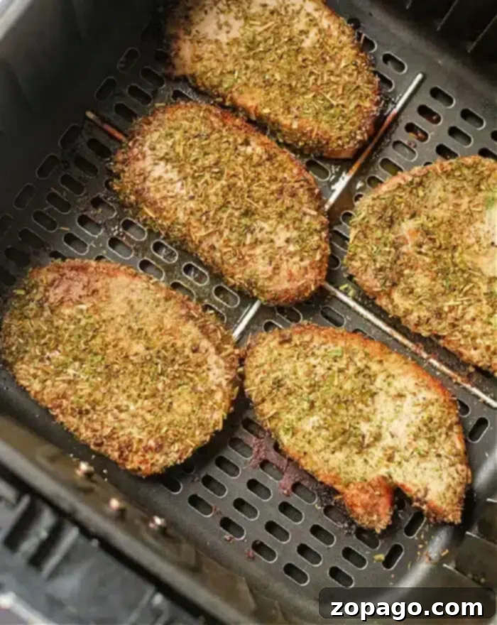 Perfect Air Fryer Pork Steak 2 Five seasoned pork steaks are evenly spaced in an air fryer basket. They are covered in a mixture of herbs and spices, giving them a crispy, golden-brown appearance.