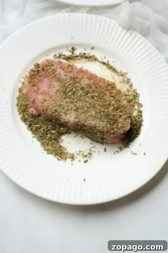 Perfect Air Fryer Pork Steak 4 A pork steak, seasoned with a variety of herbs, rests on a white, ridged plate. The finely chopped herbs blanket the top of the meat and spill over onto the plate. Perfect for an air fryer, the background sets a light, textured scene.