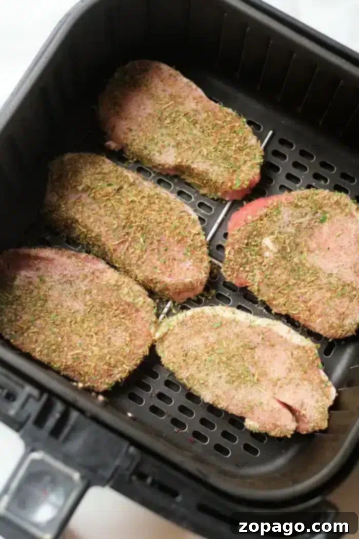 Perfect Air Fryer Pork Steak 5 Five uncooked, seasoned pork steaks are placed in an air fryer basket, ready for cooking. The meat is covered with a mixture of herbs and spices.
