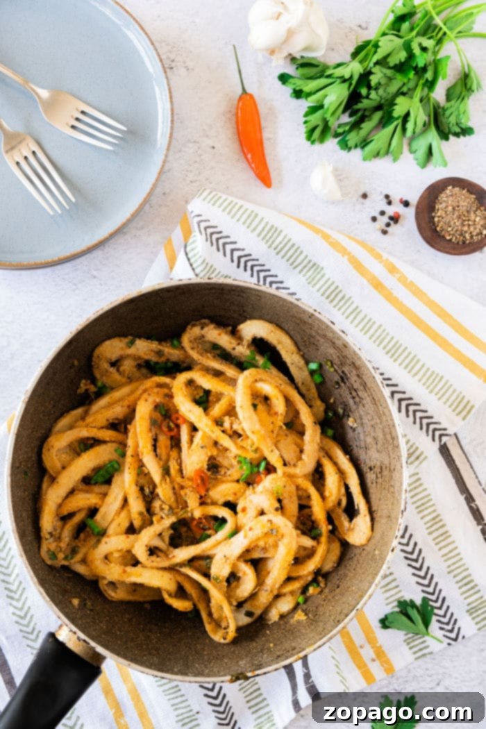 Mastering Crispy Calamari at Home 5 A pan of fried calamari with herbs and spices rests on a striped cloth beside forks, garlic, pepper, and parsley, much like an inviting setting for learning how to cook calamari.