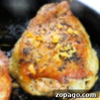 Close-up of a seasoned, golden-brown garlic butter chicken thigh in a skillet, topped with minced garlic and herbs.