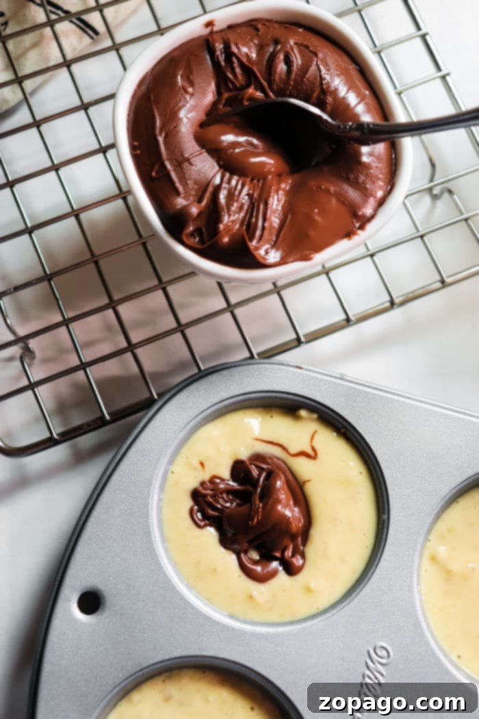 Nutella-Swirled Banana Muffins 5 Muffin batter meticulously portioned into a pan, each cup receiving a generous dollop of rich chocolate, prepared to transform into irresistible banana Nutella muffins. Next to it, a bowl of melted chocolate hints at the delicious indulgence to come.