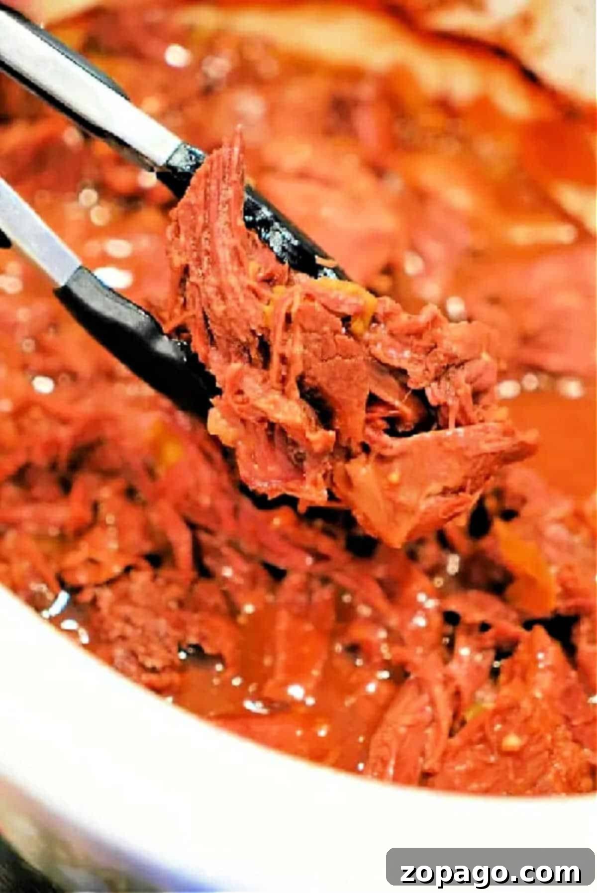 Fall Apart Slow Cooker Beef 2 Tongs holding shredded, saucy beef chuck roast over a bowl filled with more shredded beef in sauce, perfect for the slow cooker.