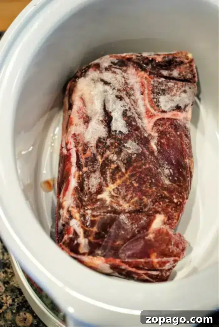 Fall Apart Slow Cooker Beef 4 A frozen beef chuck roast rests in a slow cooker, marbled with fat and covered in frost. It sits elegantly in a white ceramic dish on a granite countertop, promising tender perfection.