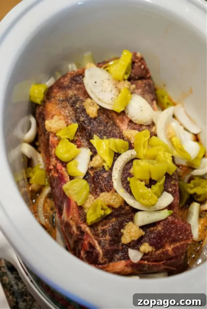Fall Apart Slow Cooker Beef 5 A seasoned beef chuck roast is placed in a slow cooker with sliced onions, garlic, and chopped yellow peppers for a flavorful dish.