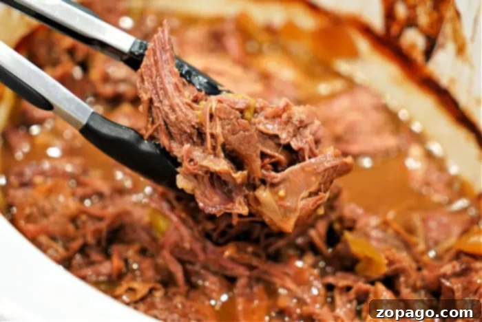 Fall Apart Slow Cooker Beef 7 Close-up of a pair of tongs holding shredded beef chuck roast over a slow cooker filled with more tender, flavorful meat covered in rich juices.