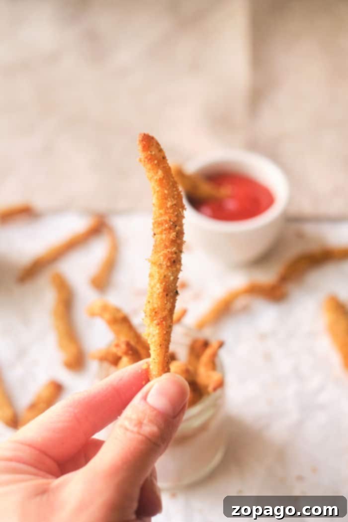 Crispy Air Fryer Chicken Fries 7 Close-up of a hand holding a crispy chicken fry, with more fries and dipping sauce in the background, reminiscent of chicken fries cooked to perfection in an air fryer.