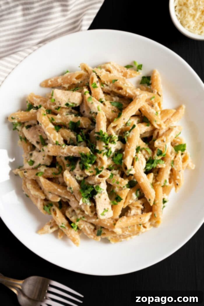 Creamy Slow Cooker Chicken Alfredo 6 A white plate filled with creamy penne pasta garnished with chopped fresh herbs. The pasta is mixed with a rich, cheesy sauce reminiscent of slow cooker chicken alfredo. In the background, there is a small bowl of grated cheese, part of a cloth, and a fork on a dark surface.