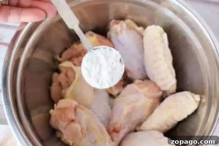 Crispy Air Fryer Chicken Wings with Baking Powder 5 A hand holds a tablespoon of baking powder, poised over a metal bowl filled with raw chicken wings, ready to be seasoned for crispy air frying.