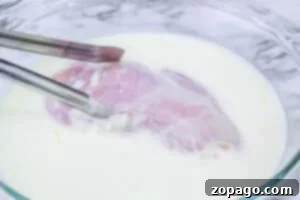 Unlock the Secret McChicken Recipe 6 Raw chicken breast being dipped in a bowl of milk with metal tongs on a marble surface, the first step to recreating an Air Fryer McDonalds Fried Chicken Sandwich at home.