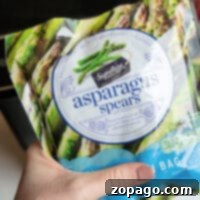A hand holds a package of Signature Select frozen asparagus spears, perfect for the air fryer. The front label features images of asparagus and highlights product features such as being steamable and resealable.