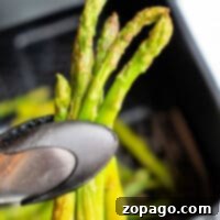 A close-up of a tong holding perfectly cooked, crispy asparagus spears over an air fryer basket, ready to serve.