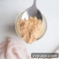 Homemade Lipton French Onion Soup Mix 7 A spoon holds a scoop of tan, flaky French Onion Soup Mix powder over a white surface, with a soft pink cloth draped nearby.