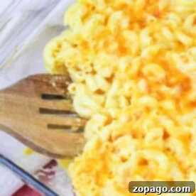 A clear glass baking dish filled with copycat Chick Fil A mac and cheese, topped with melted and lightly browned cheese. A wooden slotted spatula rests in the dish, partially lifting a portion of the macaroni. A red and white striped cloth is underneath the dish.