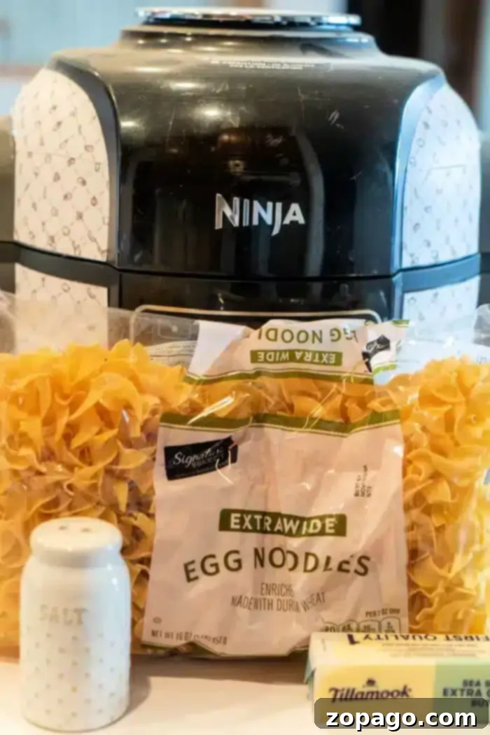 Ninja Foodi Garlic Butter Noodles 4 A sleek black Ninja Foodi air fryer looms in the background. Front and center, you'll find packages of egg noodles, a block of creamy Tillamook butter, and a chic white salt shaker adorned with gold dots—perfect ingredients for crafting delightful butter noodles.