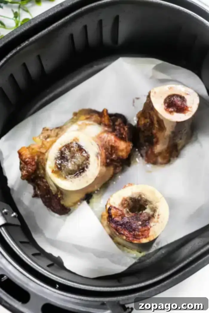 Three pieces of perfectly roasted air fryer bone marrow sit on parchment paper in an air fryer basket, showing golden and slightly charred surfaces.