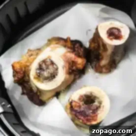 Three pieces of roasted air fryer bone marrow sit perfectly on a sheet of parchment paper inside the basket, their surfaces golden and slightly charred.