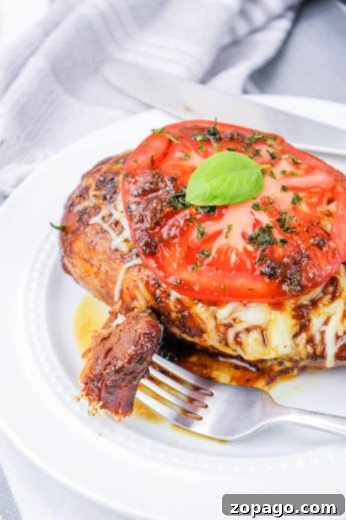Effortless Crockpot Frozen Chicken Breasts 6 Crockpot caprese chicken made from frozen breasts is topped with melted cheese, a tomato slice, and fresh basil, all elegantly arranged on a white plate with a fork and knife.