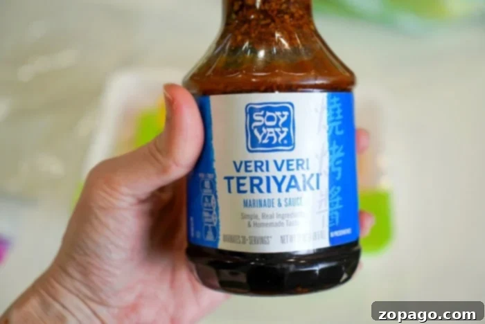 Easy Teriyaki Chicken with Bottled Sauce 4 A person holds a bottle of Soy Vay Veri Veri Teriyaki marinade and sauce, recommended for tender chicken dishes. The bottle's blue label and visible cap are featured, against a soft, out-of-focus background.
