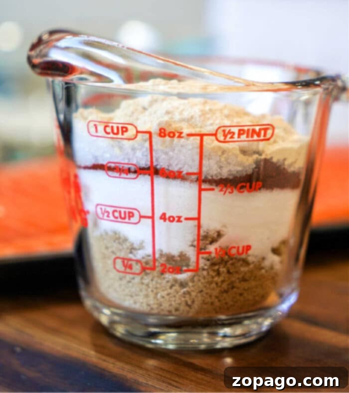 Next-Level BBQ Flavor: Homemade Dry Rub 2 A glass measuring cup on a wooden surface is filled with layers of different brown and white powders, possibly baking ingredients or components for dry rub recipes. The cup displays measurements in red from 2 to 8 ounces.