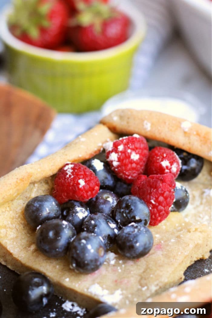 Sheet Pan Pancakes 6 A close-up of a piece of pastry topped with powdered sugar, blueberries, and raspberries. In the background, there is a green bowl filled with fresh strawberries. The scene resembles a delightful setting where oven-baked German pancakes could be served.