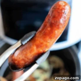 Tongs holding a perfectly cooked sausage, freshly transformed from frozen, in front of an air fryer.