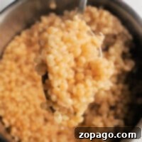 A spoon lifting cooked Israeli couscous over a bowl on a light background invites you to discover how to cook pearl couscous to perfection.