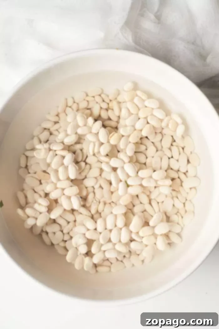 A white bowl filled with Great Northern beans submerged in water, sitting on a light-colored countertop, illustrating the soaking process.
