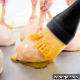 Basting lollipop chicken drumsticks with oil on a baking sheet using a brush.