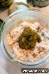 A glass bowl brimming with a creamy blend, perhaps reminiscent of a tuna melt, adorned with chopped pickles. A silver spoon peeks from the side, while jar lids linger in the background.