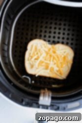 A slice of bread topped with shredded cheese and tuna sits inside the air fryer basket. The air fryer is open, revealing the perforated surface beneath this delectable Air Fryer Tuna Melt.