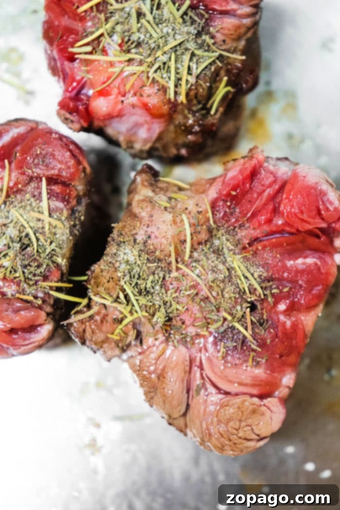 Three raw pieces of oxtails, seasoned with herbs and spices on a metal surface. The herbs are visibly sprinkled on top, adding texture and color contrast to the red and pink hues of the meat.