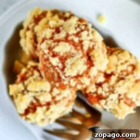 A plate contains three crumb-topped mini pumpkin muffins, showcasing a golden-brown streusel layer. A fork with a wooden handle is partially visible next to the muffins, suggesting readiness for eating. The background is blurred, emphasizing the food as the main focus.