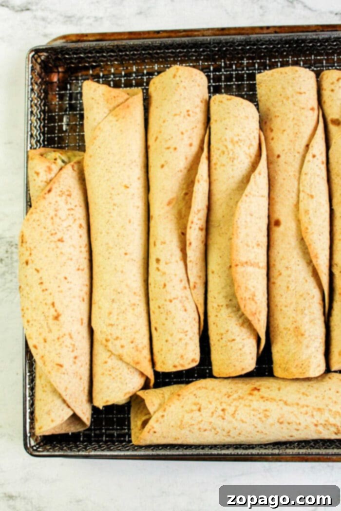 Homemade Crispy Air Fryer Chicken Taquitos 6 A tray of neatly arranged chicken taquitos rolled to perfection sits on a metal rack. The tortillas, cooked evenly in an air fryer, are light brown with visible specks. Beneath the tray lies a white, textured surface.