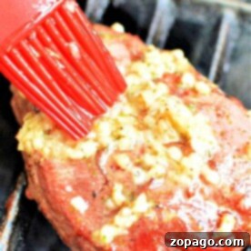 A red brush applies garlic butter to a piece of frozen steak on the grill, illustrating how to grill frozen steak perfectly.