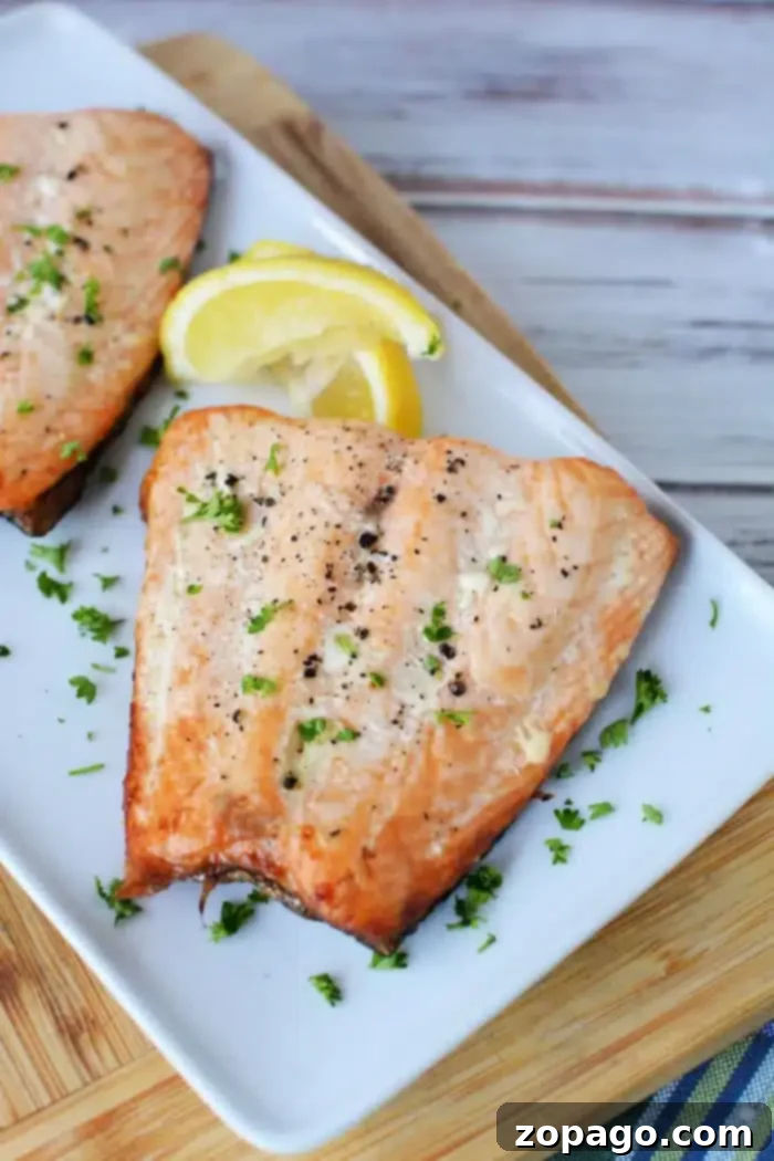 How to Grill Salmon from Frozen 2 A delectable grilled salmon fillet rests on a rectangular white plate, garnished with chopped parsley and black pepper. Two lemon wedges accompany this delight. The plate, showcasing perfectly thawed frozen salmon grill markings, sits on a wooden cutting board with a striped cloth beneath.