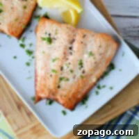 A grilled salmon fillet, previously frozen for freshness, glistens with chopped parsley on a white plate. Lemon wedges accompany it, all elegantly positioned on a wooden cutting board atop a plaid fabric.