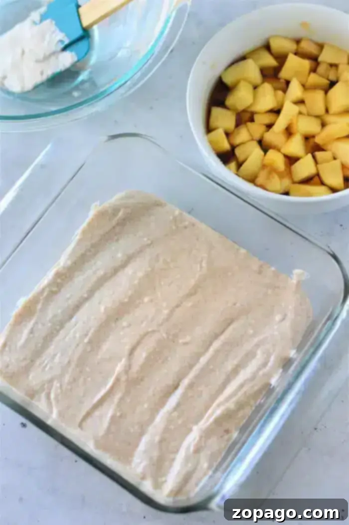 A glass baking dish holds a layer of batter spread evenly. Beside it are two bowls: one with chopped apple pieces and another with a blue spatula resting on the edge, containing a white creamy mixture. This Apple Cobbler with Bisquick is set against a light-colored countertop background.