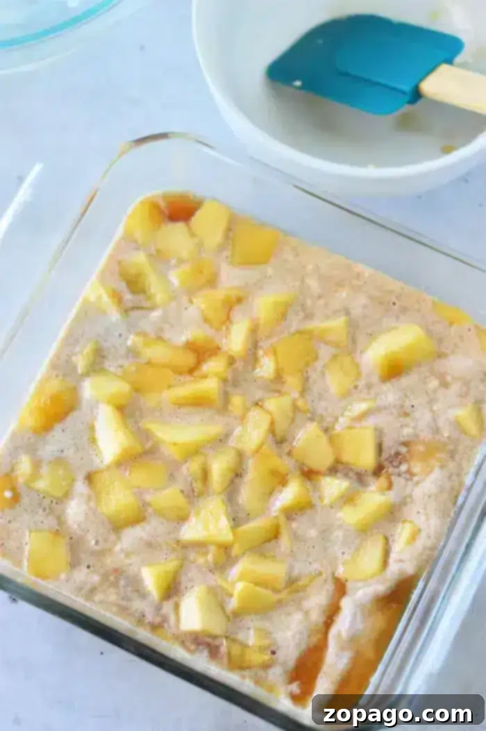 Easy Bisquick Apple Cobbler 7 A clear square baking dish filled with a mixture of diced apples and Bisquick batter. A blue spatula rests inside a white mixing bowl in the background, ready to complete this delicious Apple Cobbler with Bisquick.