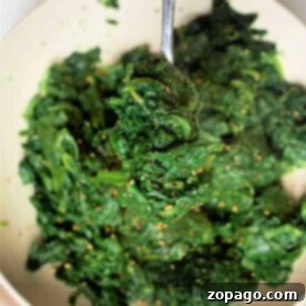 A hand holds a fork with cooked spinach seasoned with spices on a white plate, offering inspiration for those wondering how to cook frozen spinach deliciously.