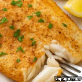 Golden, pan-seared fish fillet made from Frozen Tilapia Air Fryer, garnished with parsley and served with lemon wedges and a forkful of fish.