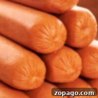 A close-up image reveals a stack of smooth, orange-colored hot dog sausages, their ends sealed and slightly pinched. Tightly packed to showcase their glossy texture and cylindrical shape, these sausages prompt the question: how long to boil hot dogs for perfect tenderness?